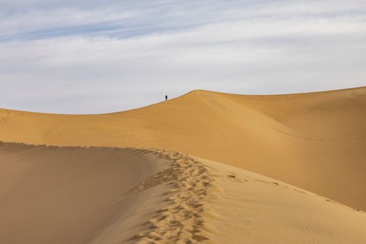 A lone hiker walks across vast sand dunes under a blue sky, showcasing nature's beauty.