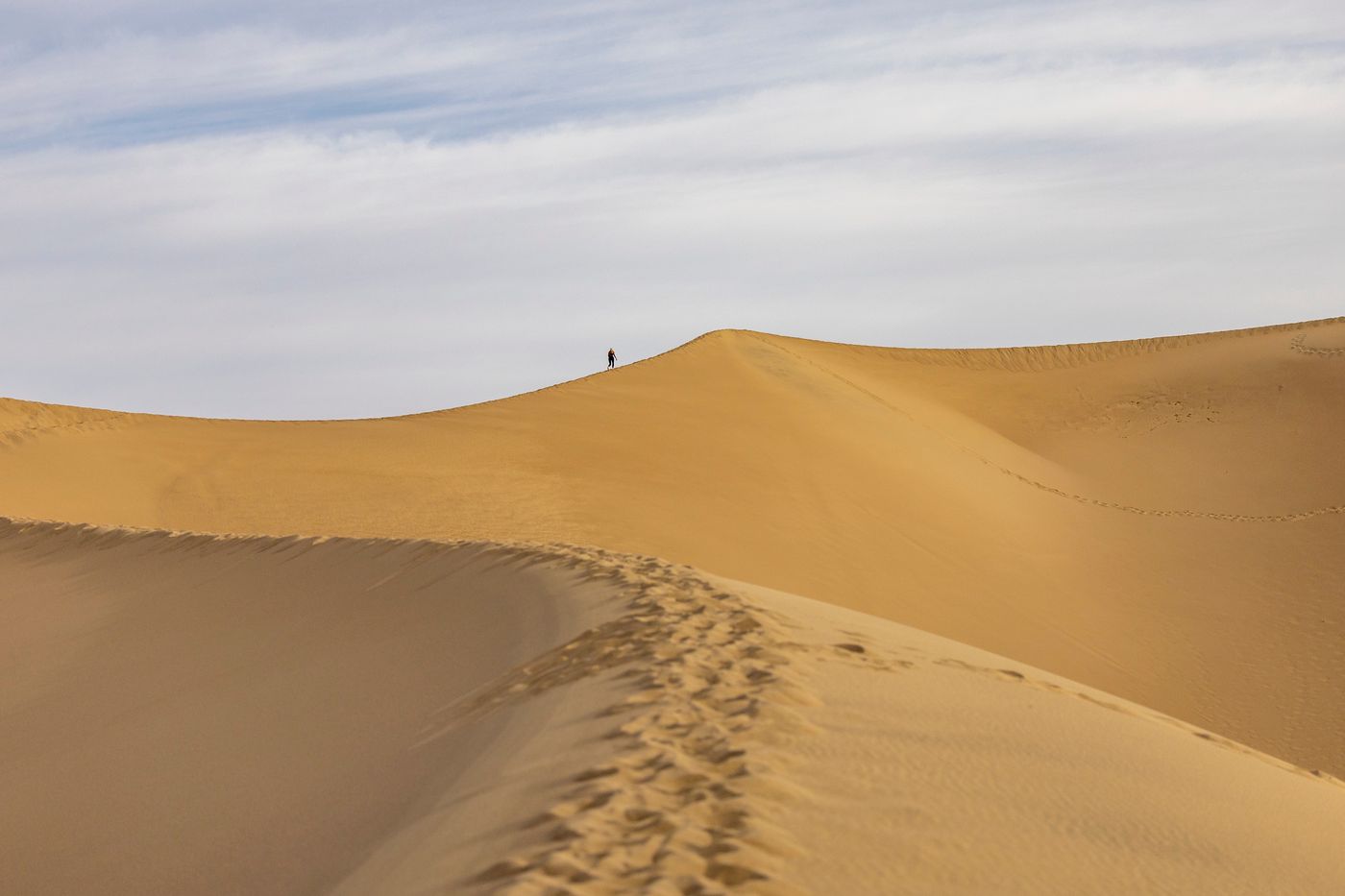 A lone hiker crossing vast sand dunes under a blue sky by Stephen Leonardi on Pexels