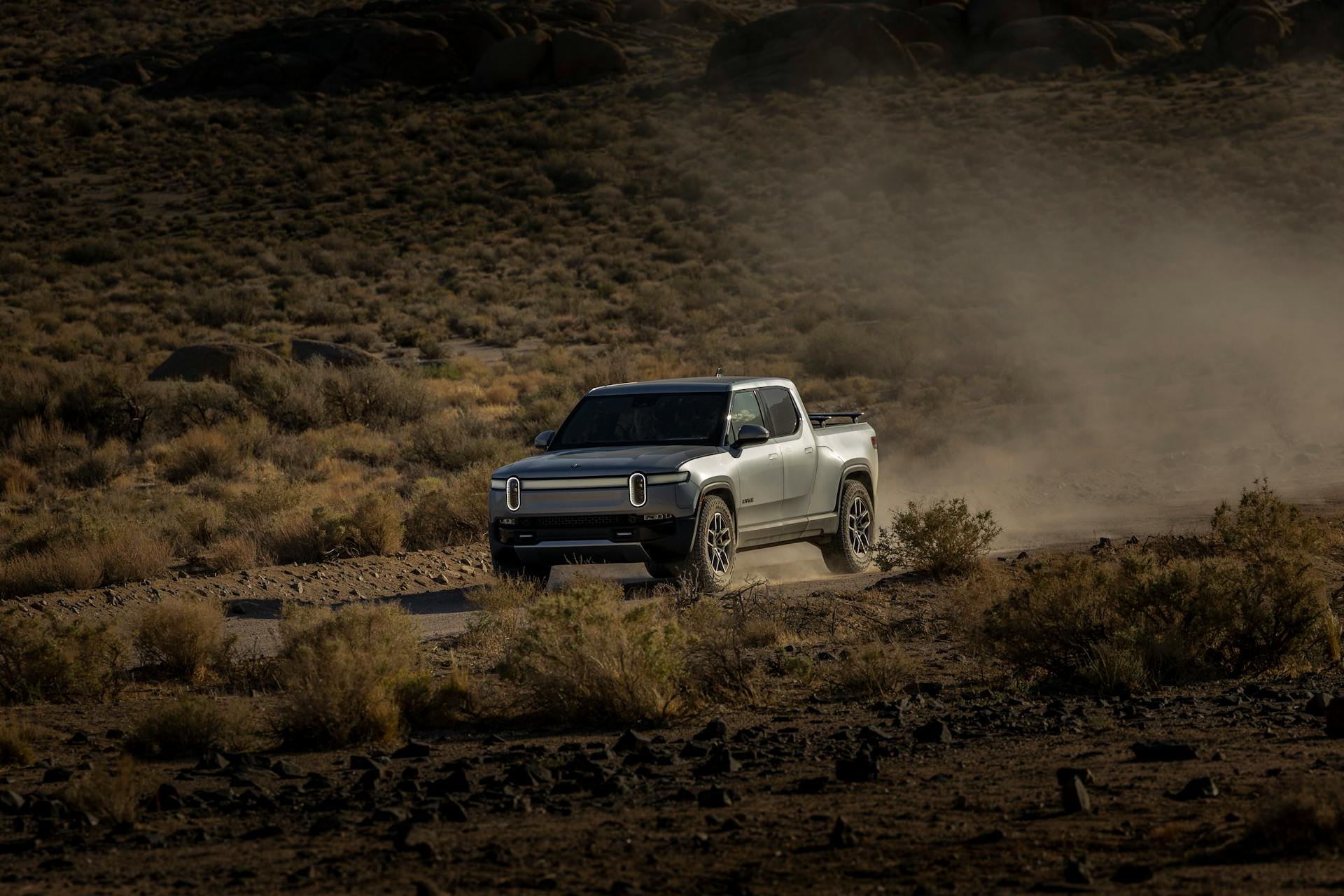 Modern electric truck navigating rugged desert terrain, raising dust clouds.