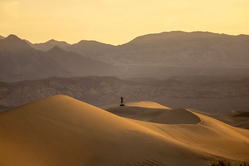 Free Person stands on vast desert dunes at golden hour with mountains in the background. Stock Photo