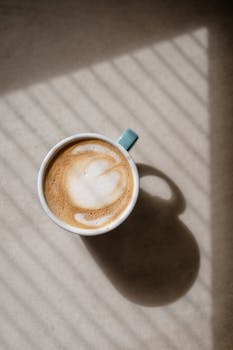 Cappuccino with latte art in sunlight through blinds, creating soft shadows.