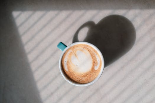 A top-down view of a cappuccino with heart latte art, casting a shadow in morning sunlight.