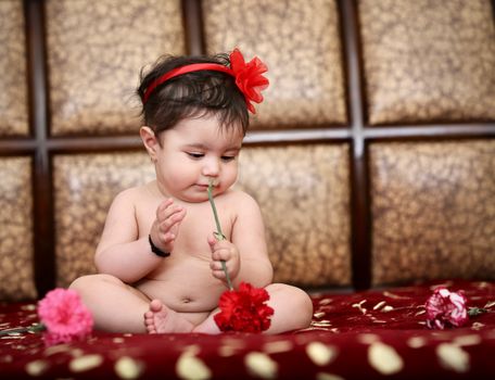 Adorable baby with red flower headband holding a red carnation indoors.