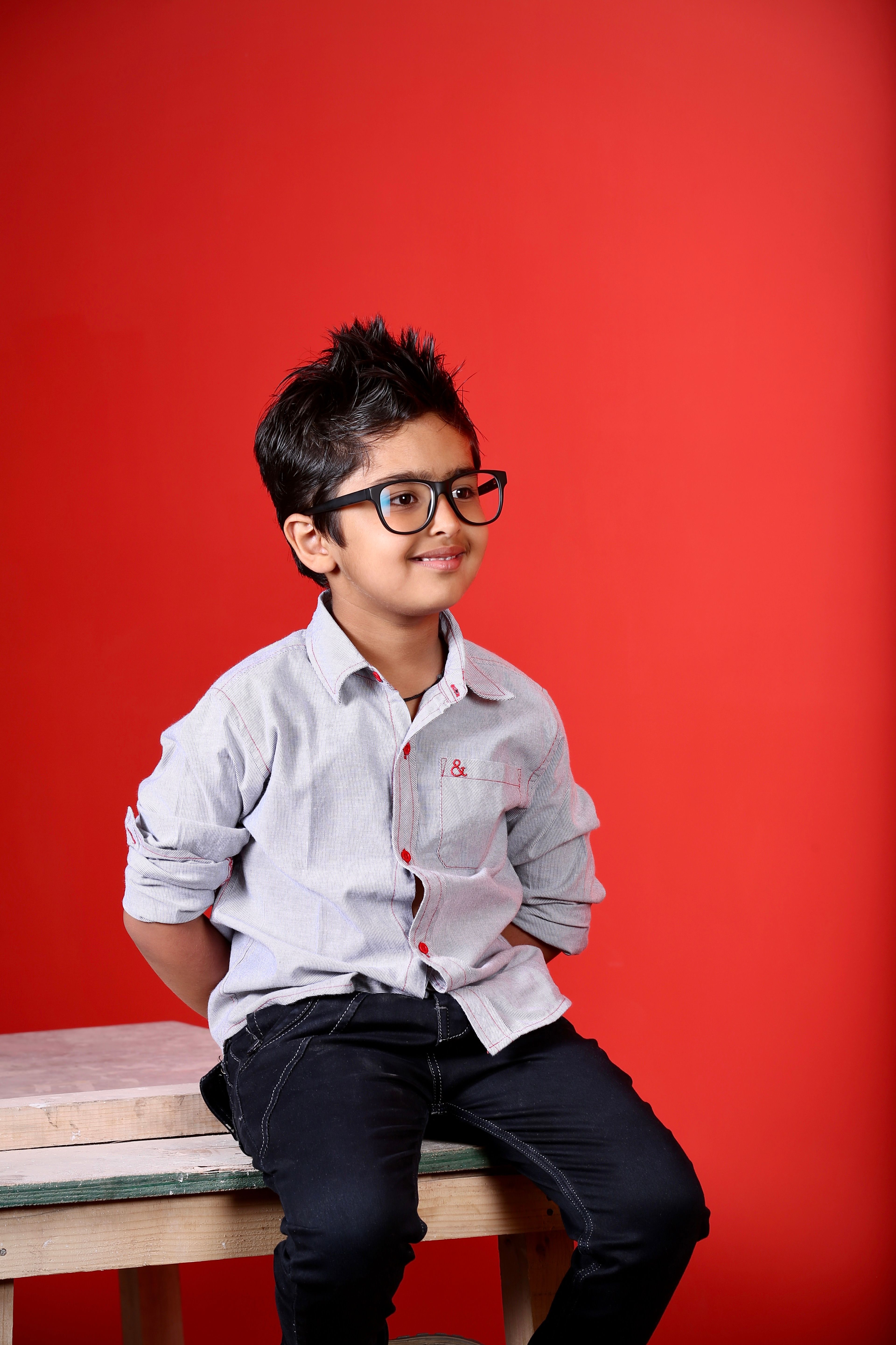 Cute young boy wearing glasses seated against a vibrant red backdrop.