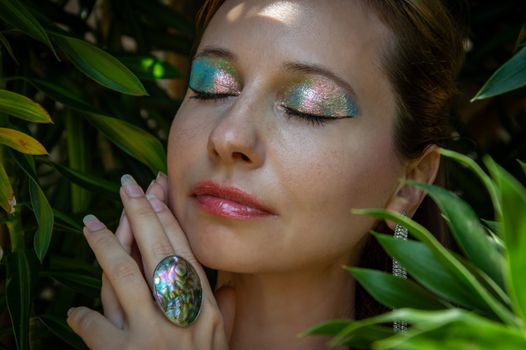 Portrait of a peaceful woman outdoors with shimmering eyeshadow surrounded by lush greenery.