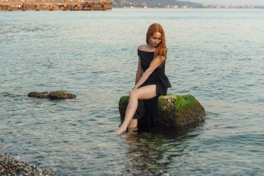 A woman in a black dress sitting on a rock by the sea, reflecting tranquility.