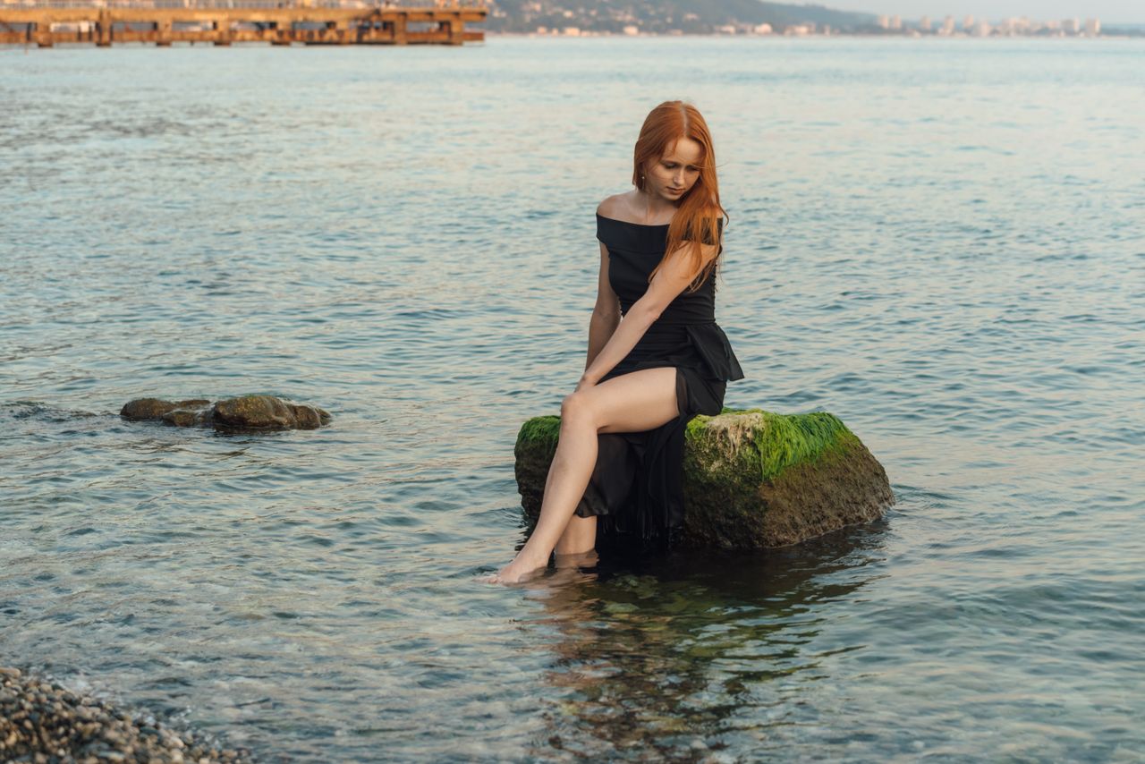 a woman in a black dress sitting on a rock by the sea reflecting tranq