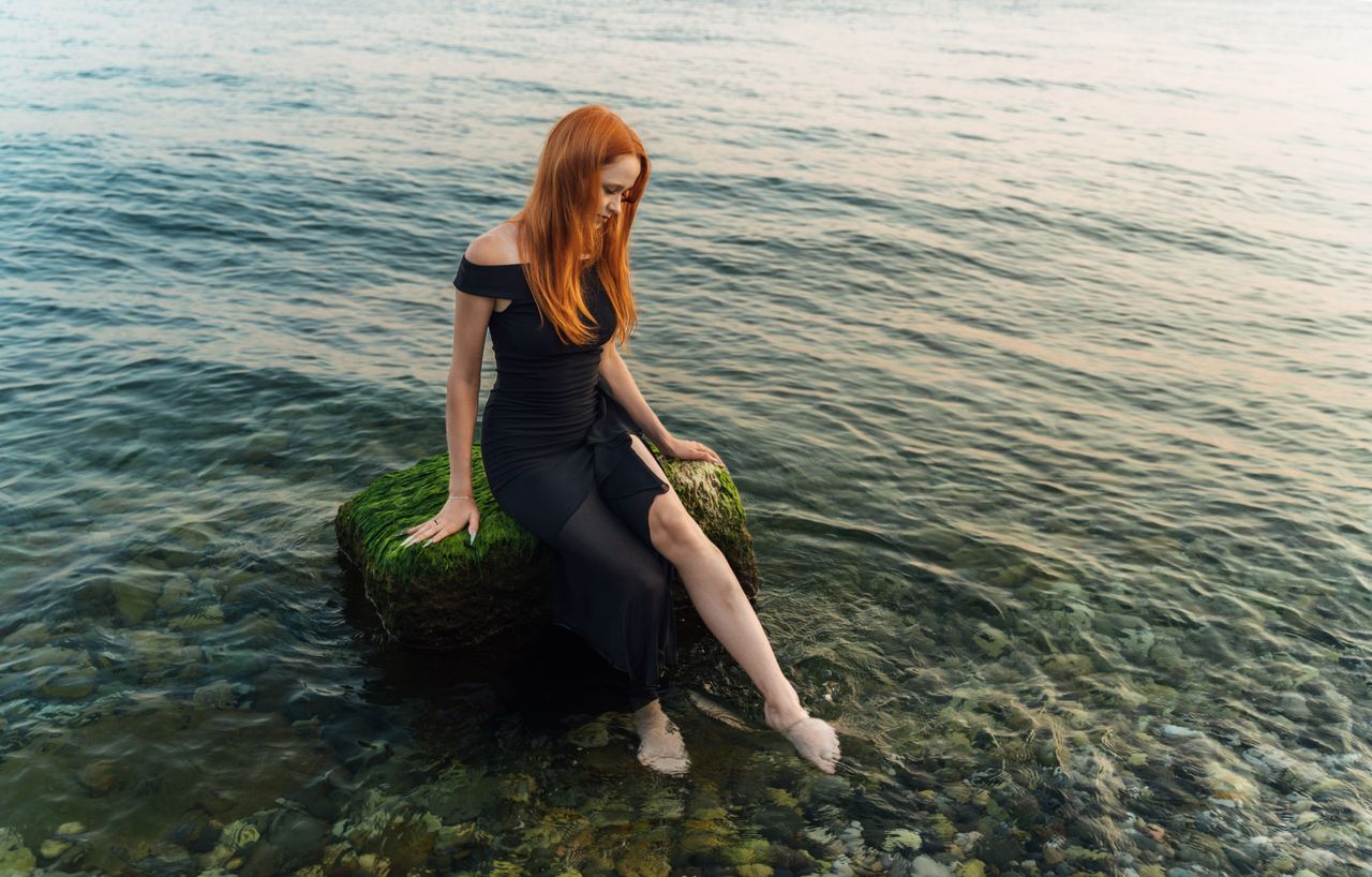 a relaxed redhead woman sits on a moss covered rock by the sea enjoyin