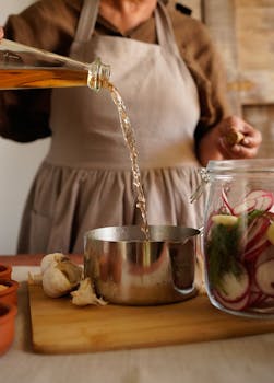 A person pours vinegar into a jar for pickling, surrounded by fresh ingredients.