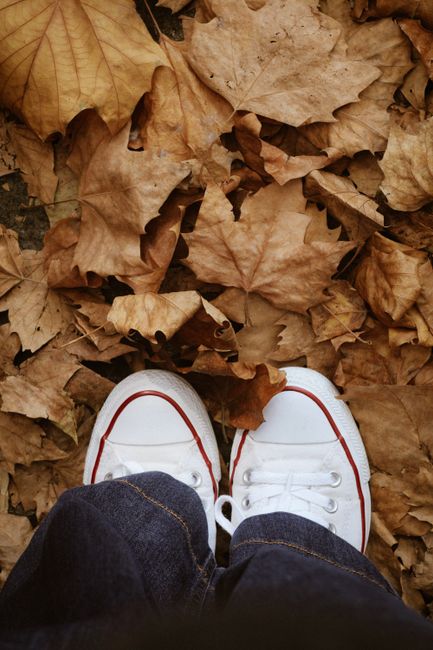 Person wearing white sneakers standing on dry autumn leaves outdoors.