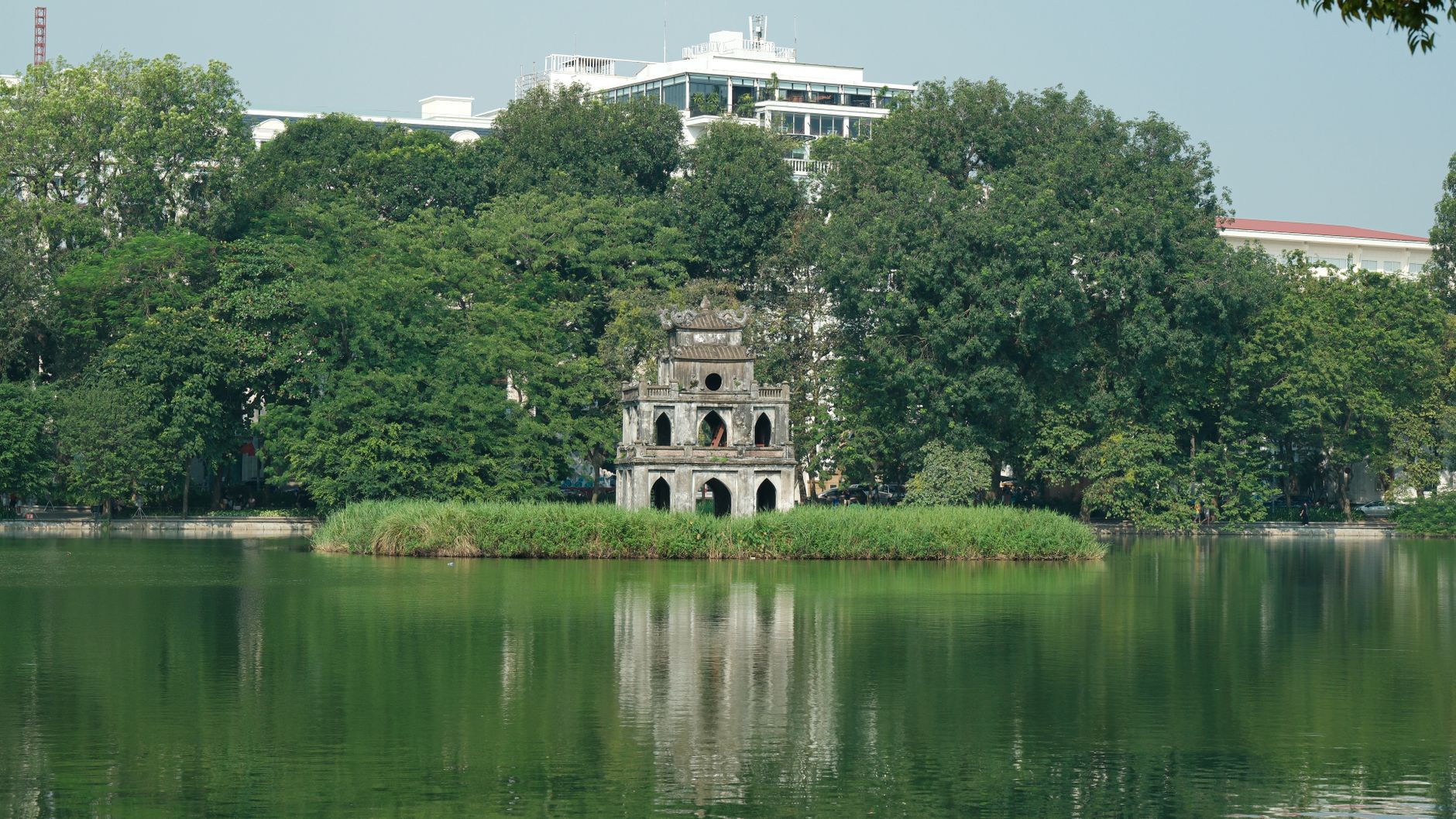 Serene scene of Turtle Tower reflecting in Hoan Kiem Lake, amidst lush greenery on a sunny day.