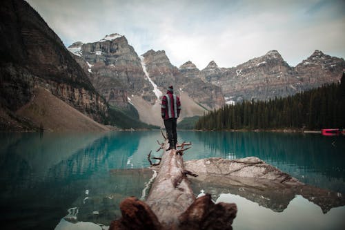 호수와 얼음 산 풍경을보고 갈색 통나무에 서있는 빨간색과 흰색 재킷을 입고 남자