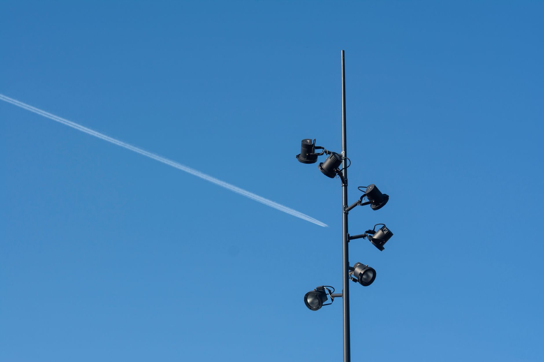 A street lamp post stands tall with a clear blue sky background and a visible contrail.