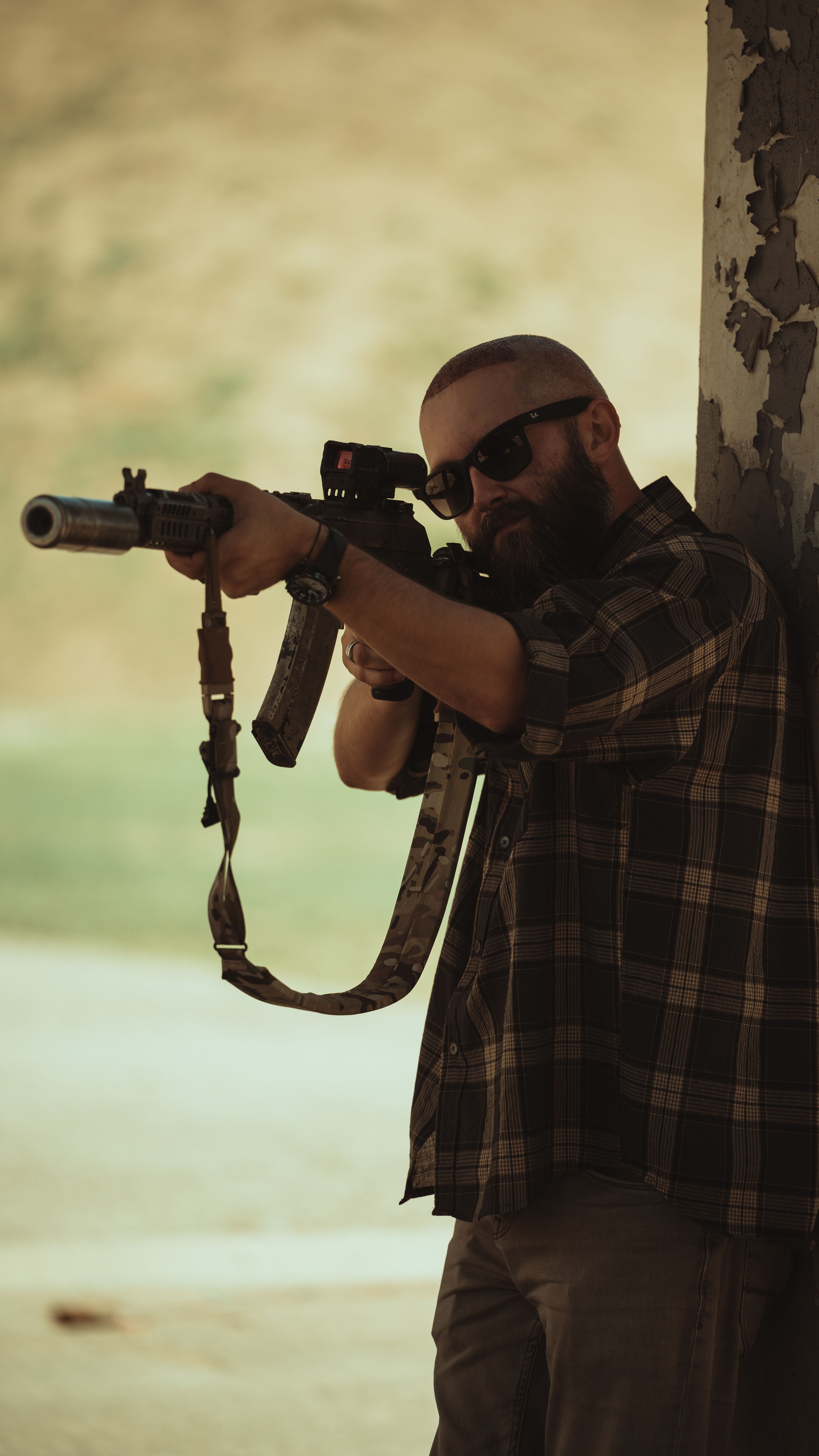 Bearded man in sunglasses aiming rifle outdoors by a worn wall.