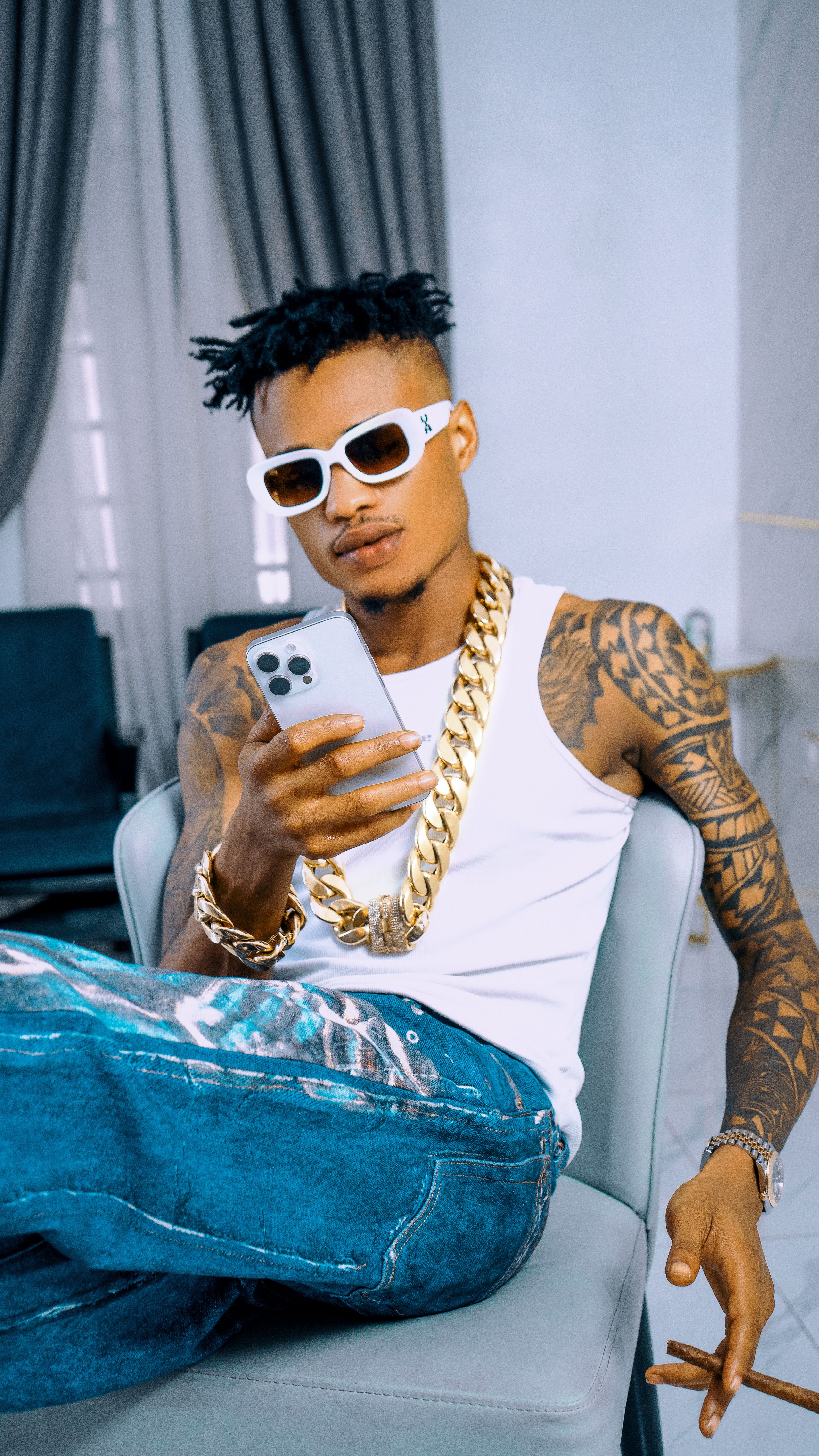 Trendy young man with tattoos and gold chains relaxing indoors with a smartphone.