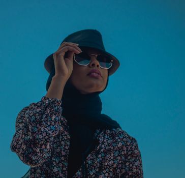 Elegant woman in a floral blouse and hat posing outdoors with sunglasses on a sunny day.