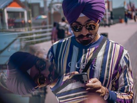 阳光明媚，一位头戴头巾、身穿条纹衬衫的时尚男子正在户外全神贯注地玩手机