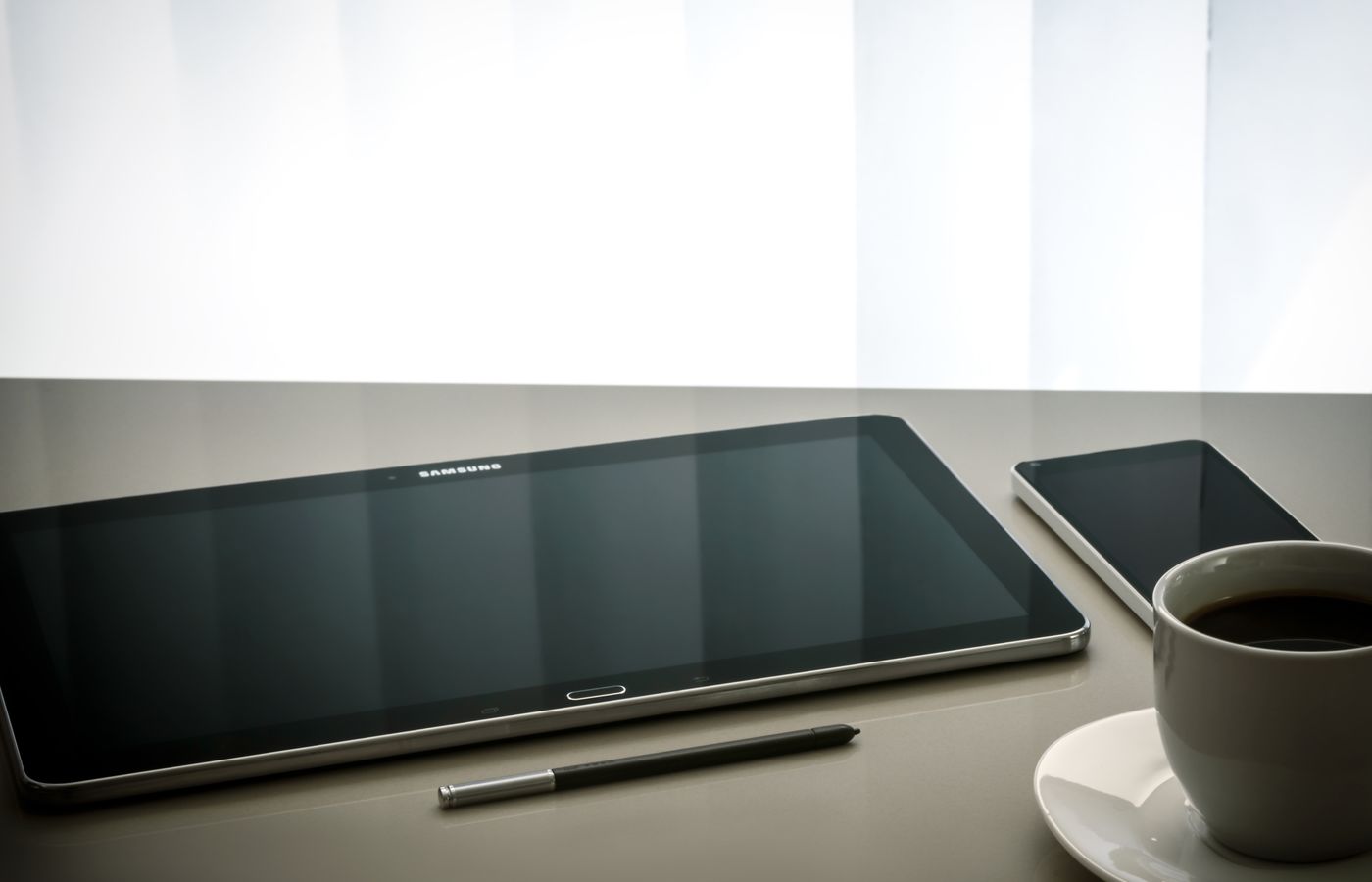 Minimalist office desk setup featuring a tablet, smartphone, pen, and coffee cup on a reflective surface.