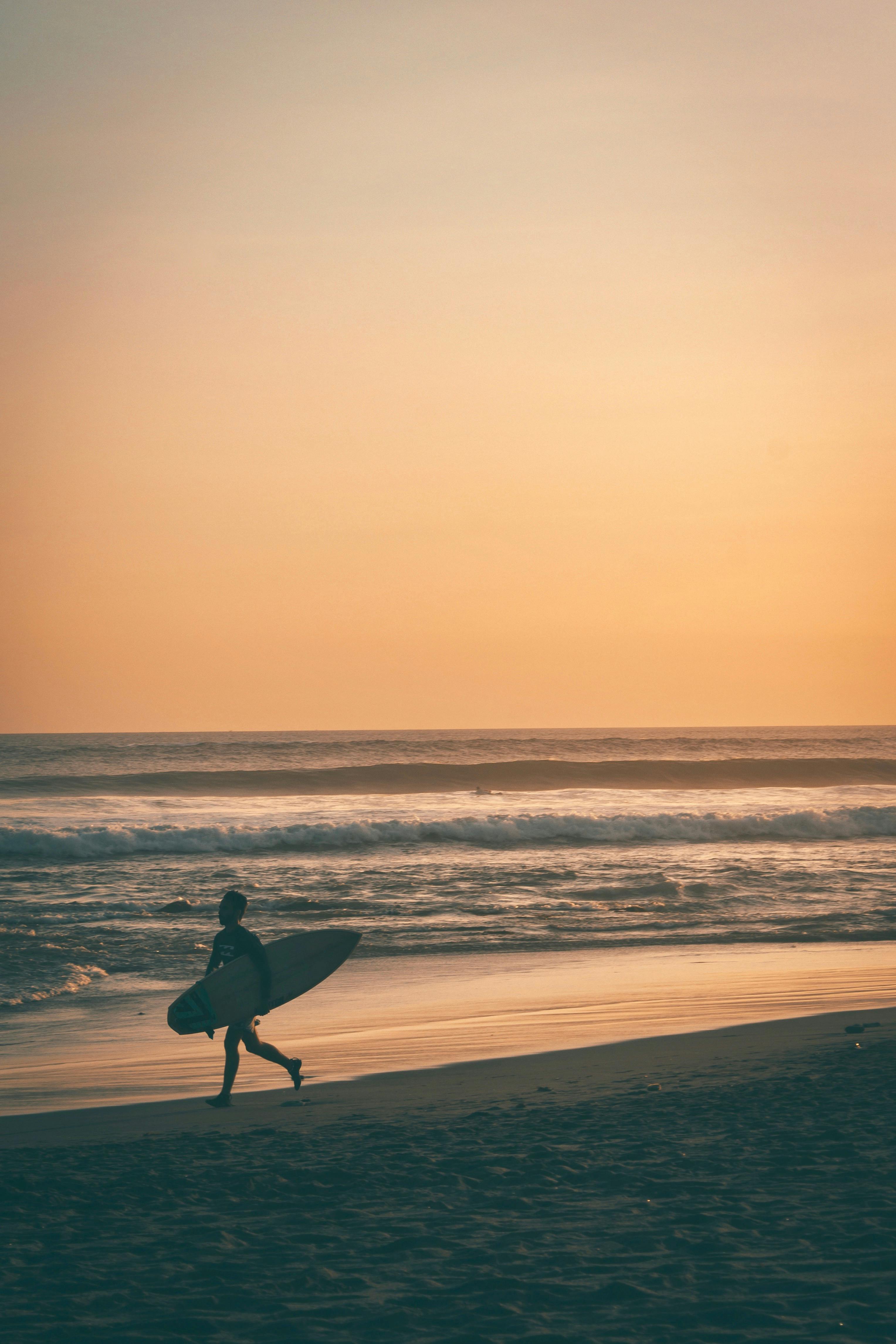 Meanwhile, a lone surfer carries a surfboard along the sandy shoreline, with gentle waves rolling in under a warm, golden sunset sky.