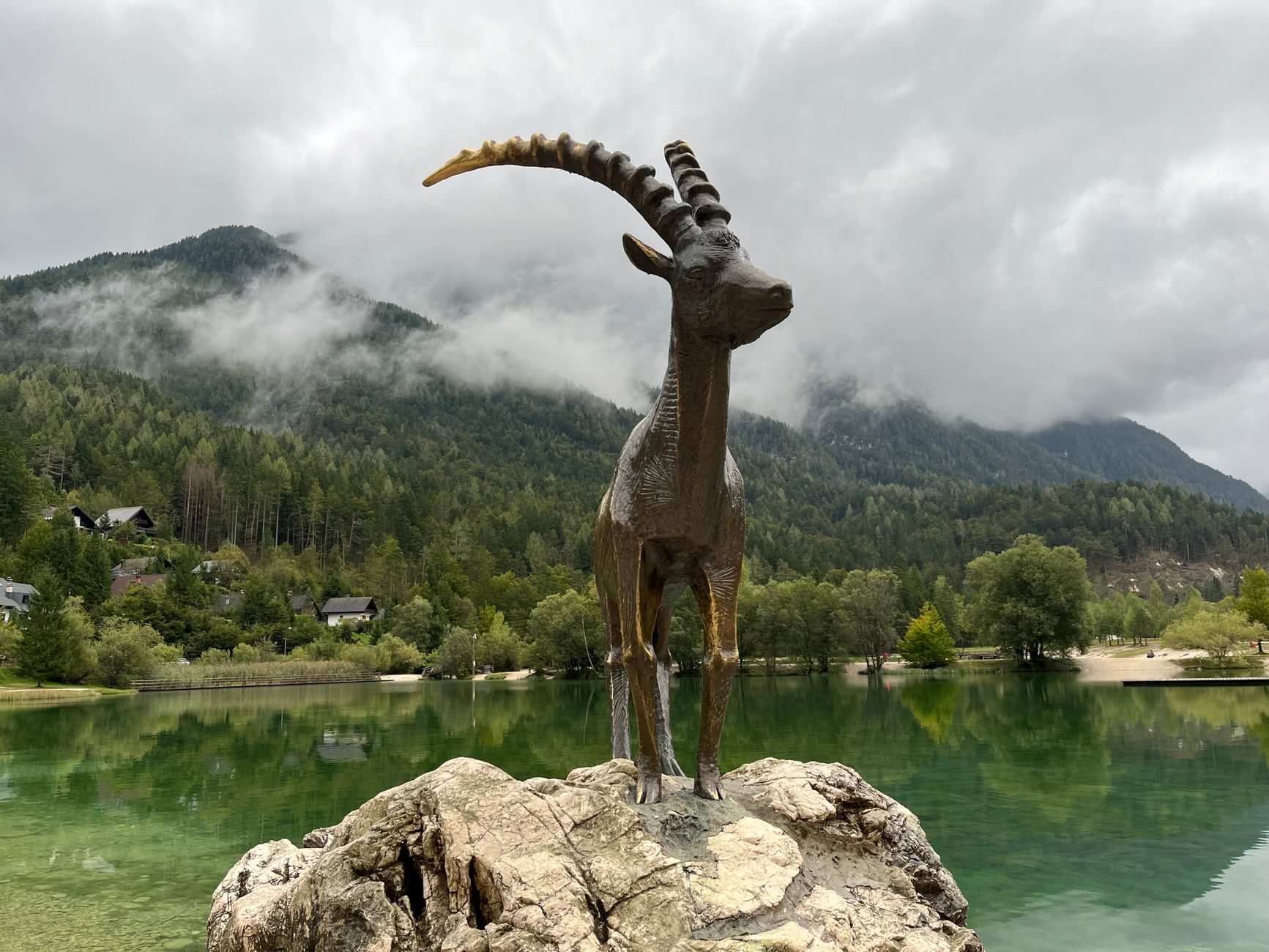 A majestic statue of an ibex on a rock by a serene Alpine lake surrounded by lush mountains.