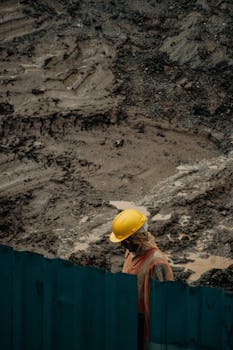 A worker with a yellow helmet stands behind a barrier at a muddy construction site.