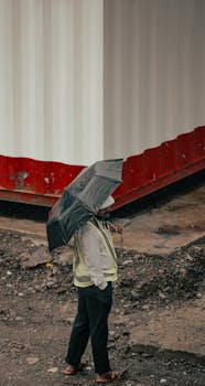 A construction worker with an umbrella walking on a rainy day beside a shipping container.