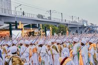 Large group of people in traditional attire participating in an outdoor religious festival near a bridge.