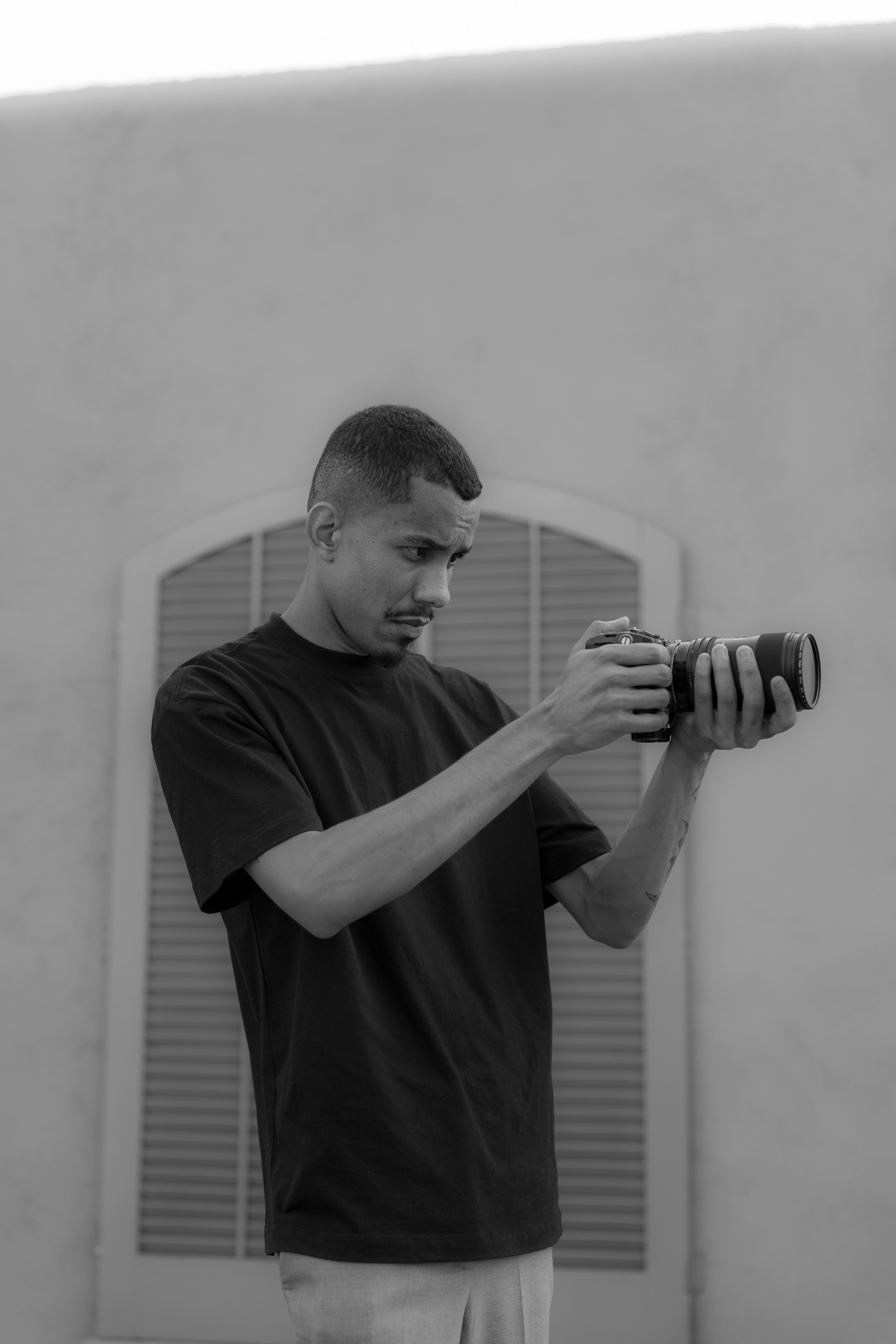 Monochrome image of a photographer analyzing camera gear against a building background.
