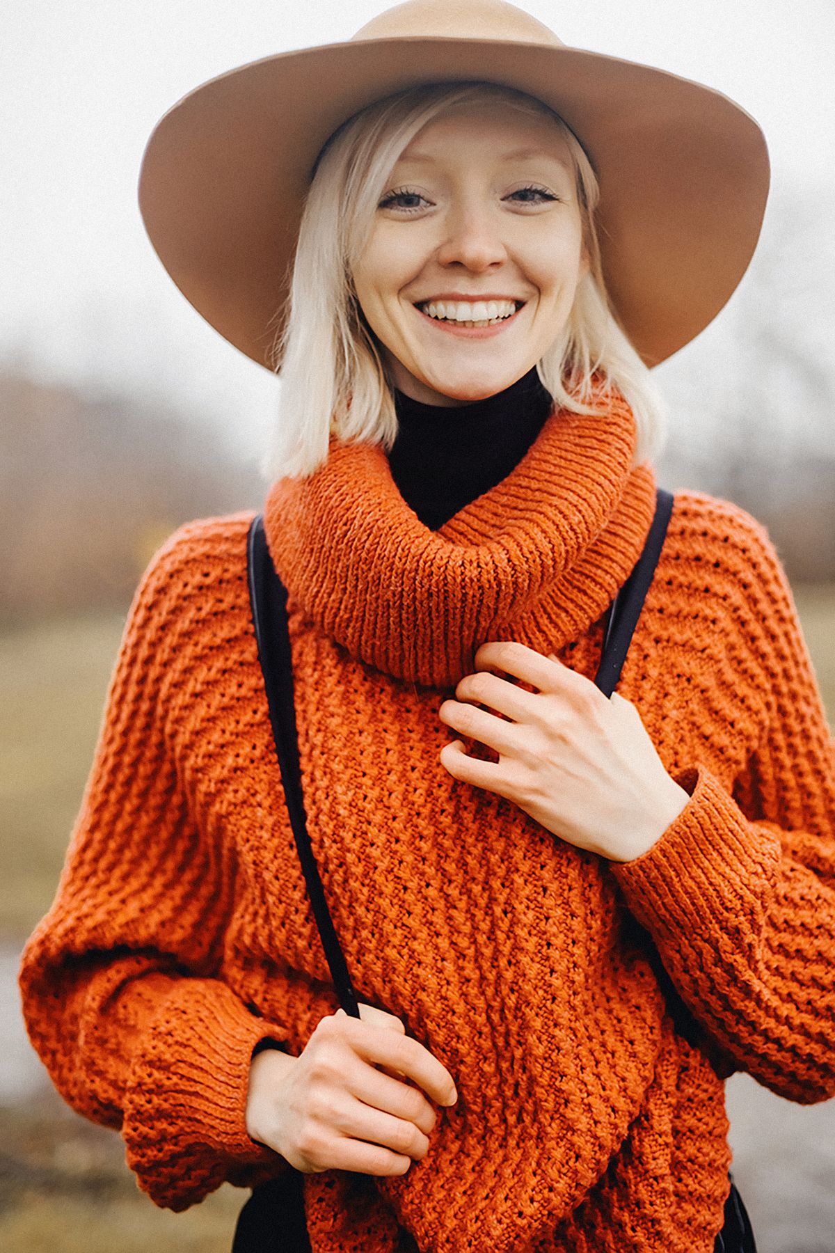 Cheerful woman wearing a cozy orange sweater and wide-brim hat outside.