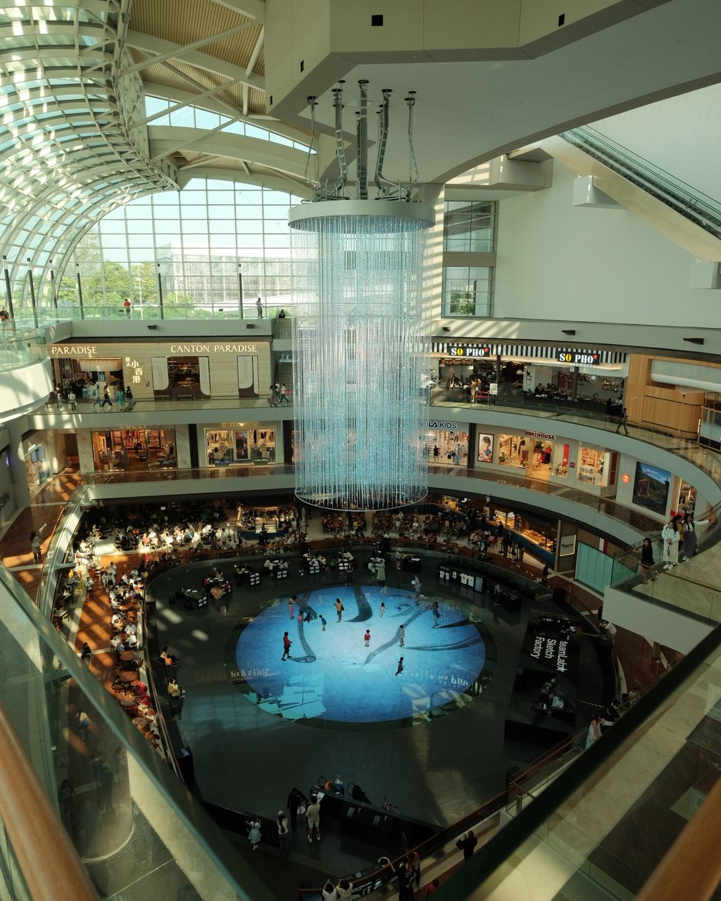 A shopping mall atrium seen from above, with a curved glass roof over a full-height void, tiered balconies of shopfronts and a long cascade of hanging lights down the centre.