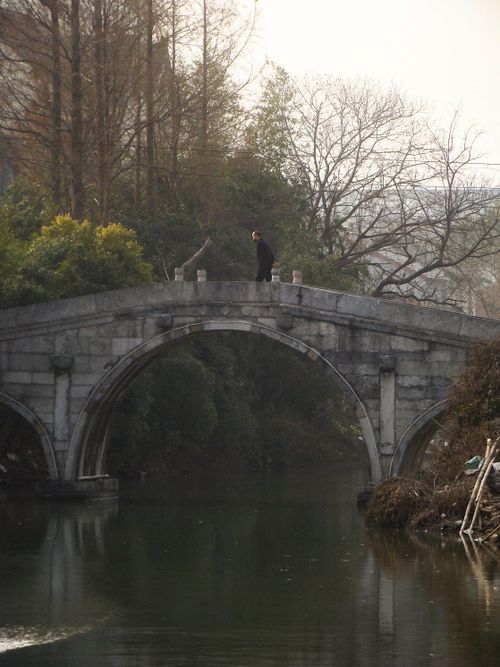 Free A serene stone bridge arches over a calm river, surrounded by lush greenery and a lone figure. Stock Photo