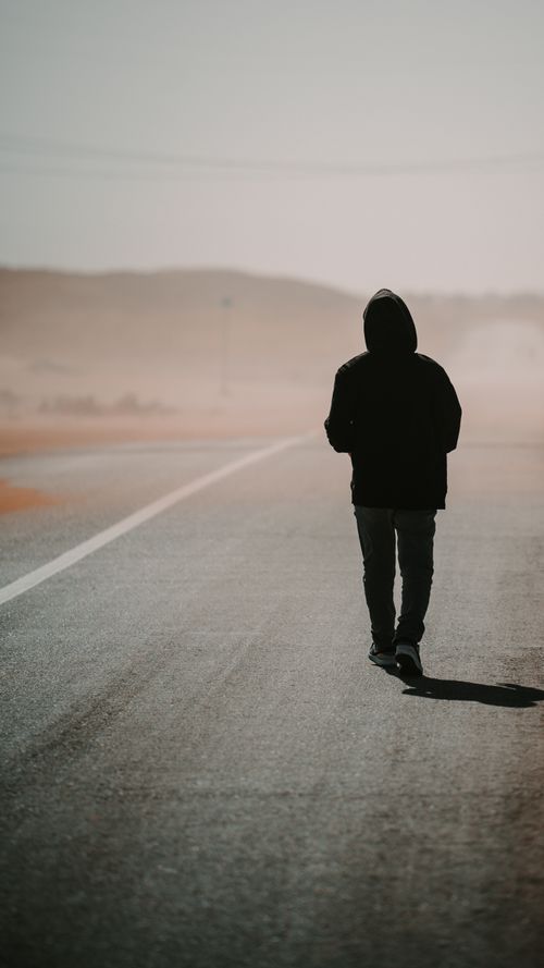 Free Hooded person walking alone on a deserted highway under a hazy sky. Stock Photo