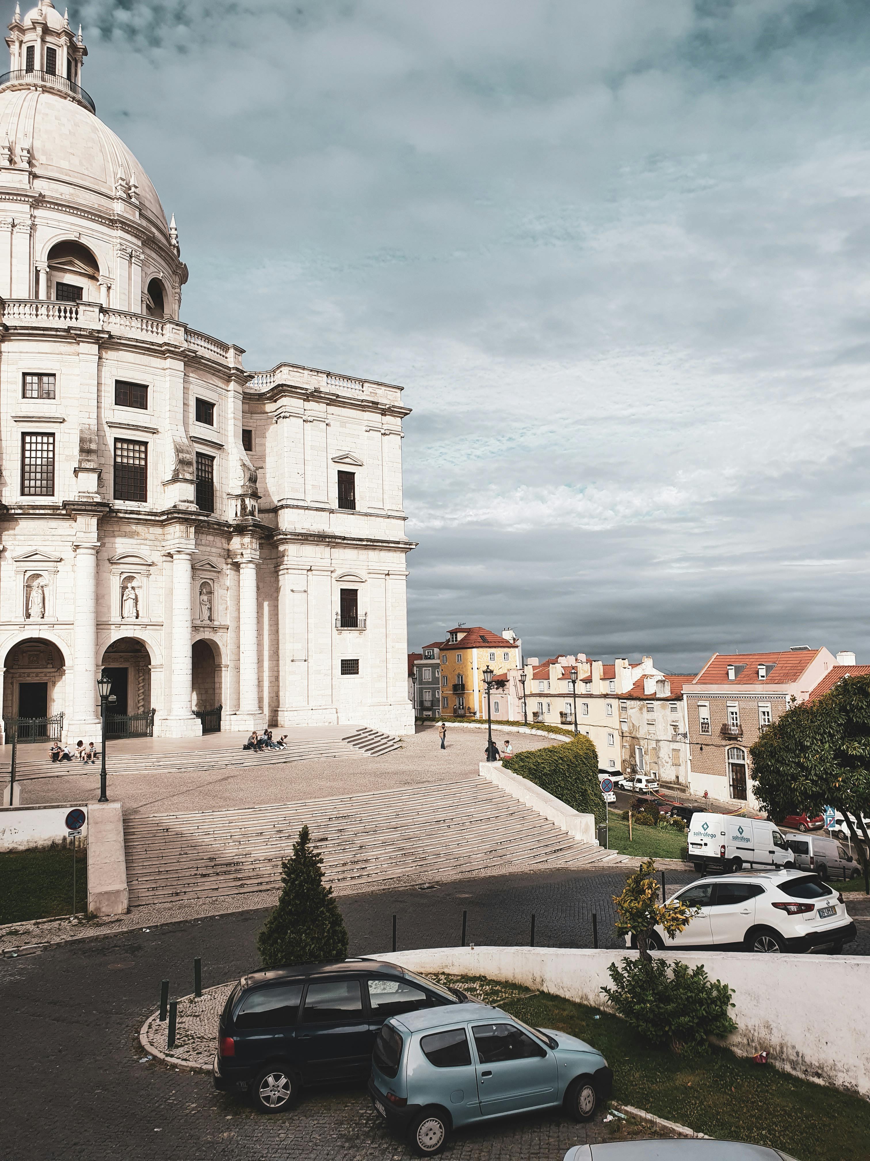 一座历史悠久的教堂，圆顶突出，以城市为背景，汽车停靠在旁，天空多云