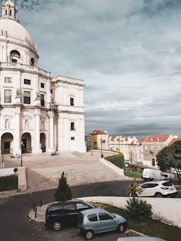 一座历史悠久的教堂，圆顶突出，以城市为背景，汽车停靠在旁，天空多云