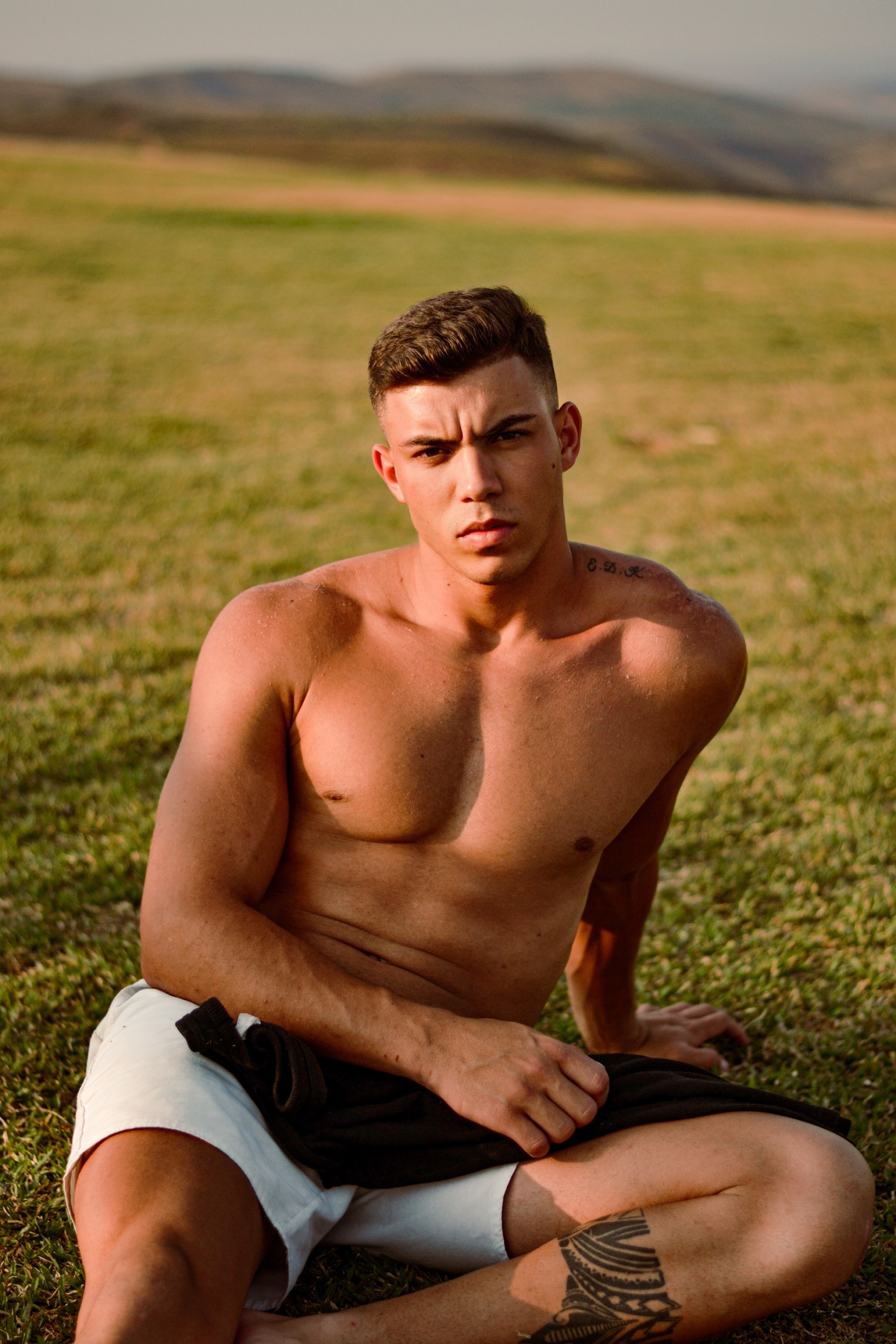 Shirtless young man with a tattoo relaxing on a grassy field under sunlight in Sete Lagoas, Brazil.
