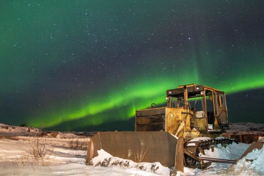 An abandoned bulldozer under the stunning aurora borealis in a snowy Arctic landscape at night.