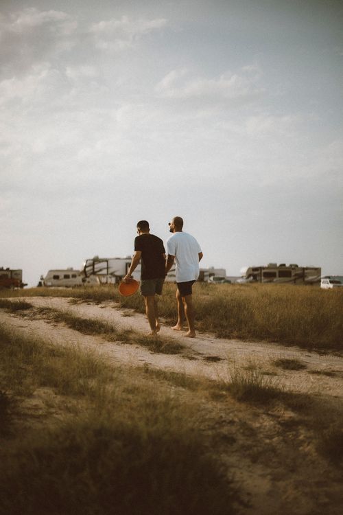 Free Two friends walking barefoot in Dubai's desert landscape, with a backdrop of parked RVs and trucks. Stock Photo