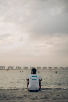 A man sits alone on a beach in Dubai, gazing at the sea under a cloudy sky.