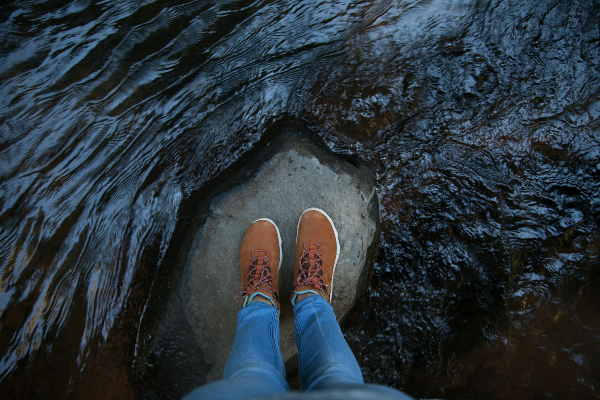 Person standing on a rock in a Sedona stream, showcasing boots and jeans, epitomizing outdoor adventure.