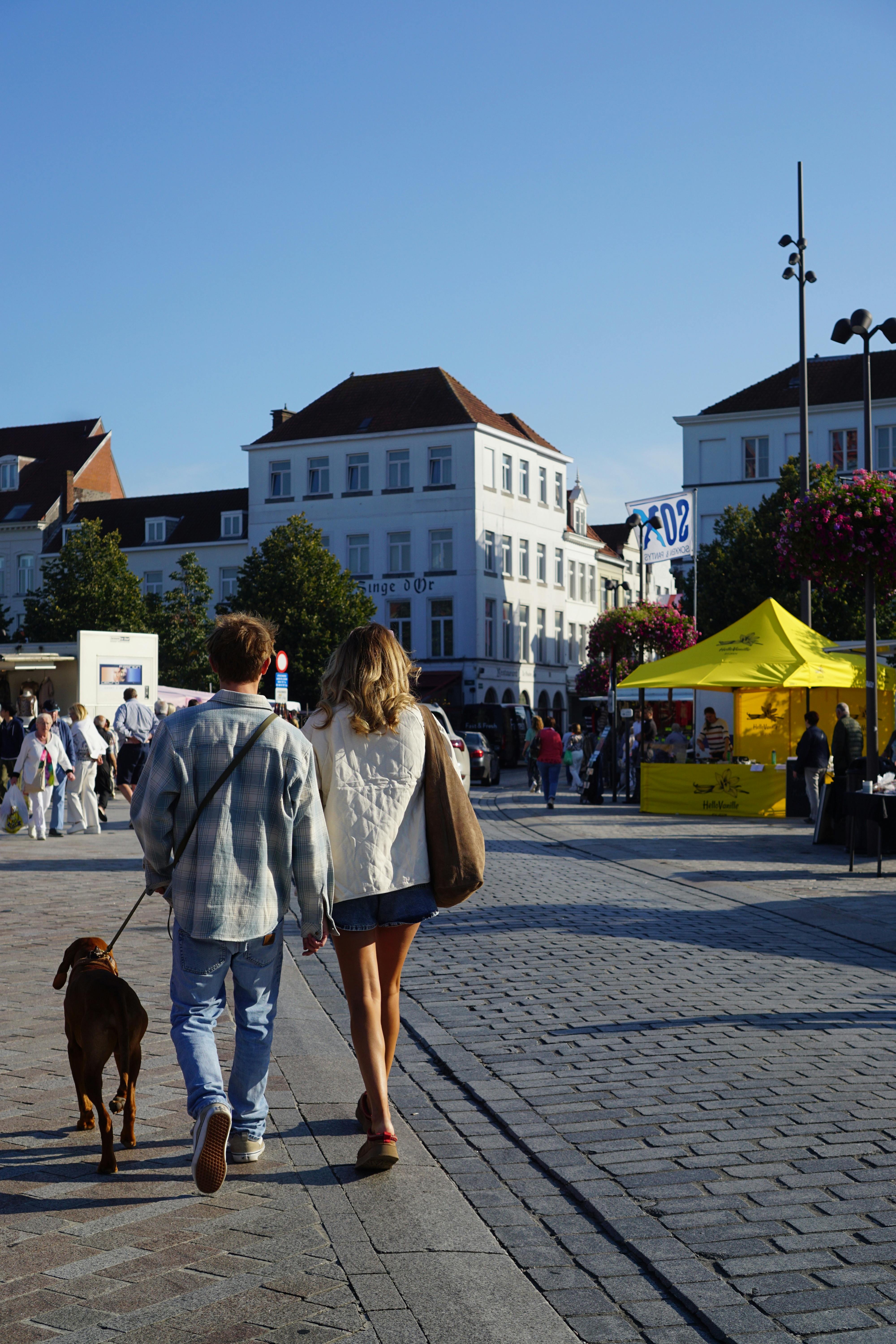Young couple walking a dog in a vibrant Bruges street market during a sunny day.
