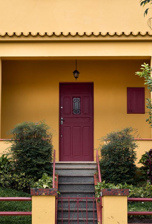 Free Charming yellow house facade with a maroon door and green shrubs accentuating the entrance. Stock Photo