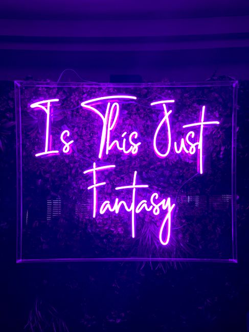 Purple neon sign with the text 'Is This Just Fantasy' glowing brightly against dark background.