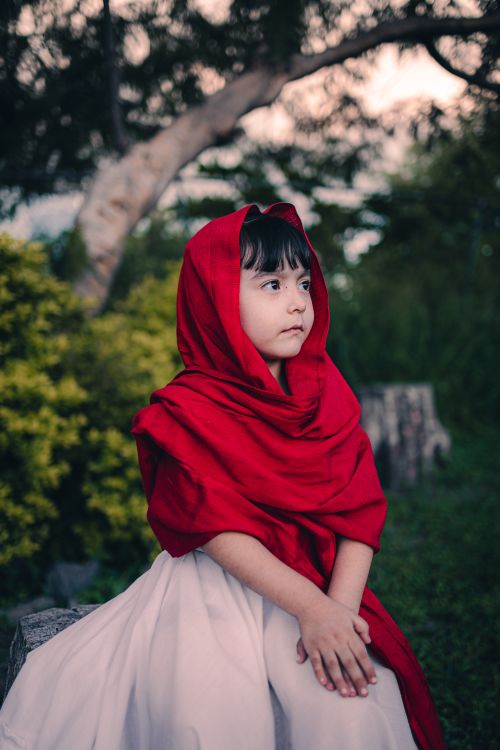 Free A young girl in a vibrant red shawl sits outdoors, exuding innocence and calm. Stock Photo