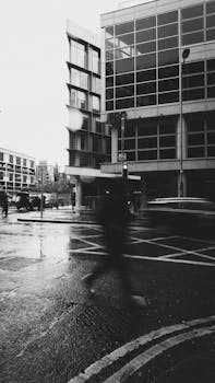 Black and white photo capturing a rainy city corner with a blurred pedestrian crossing.