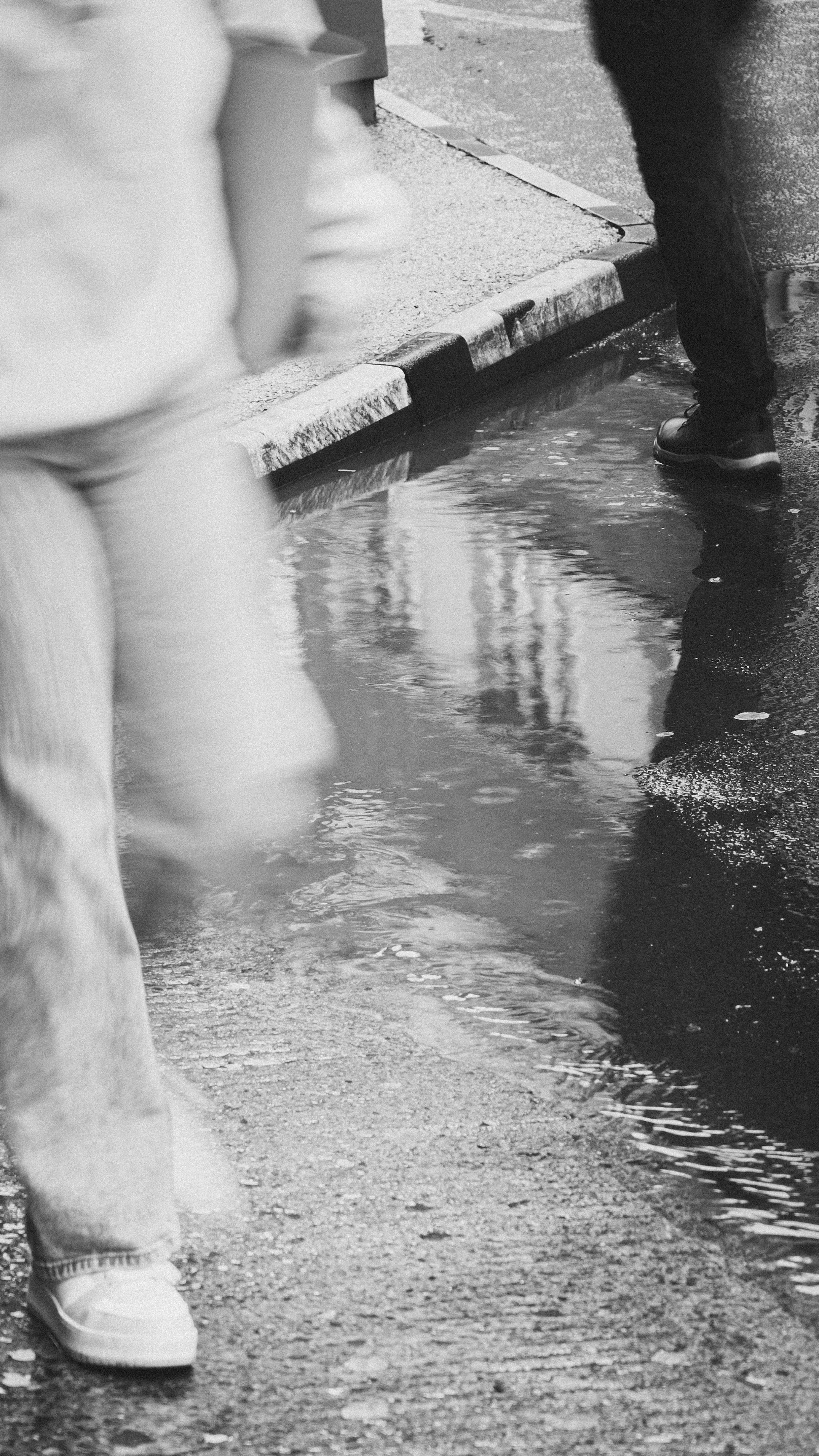 Black and white image capturing blurred walking figures by a puddle on a city street.