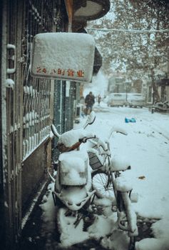 A snowy street scene featuring bicycles covered in snow, a sign with Chinese characters, and people walking.