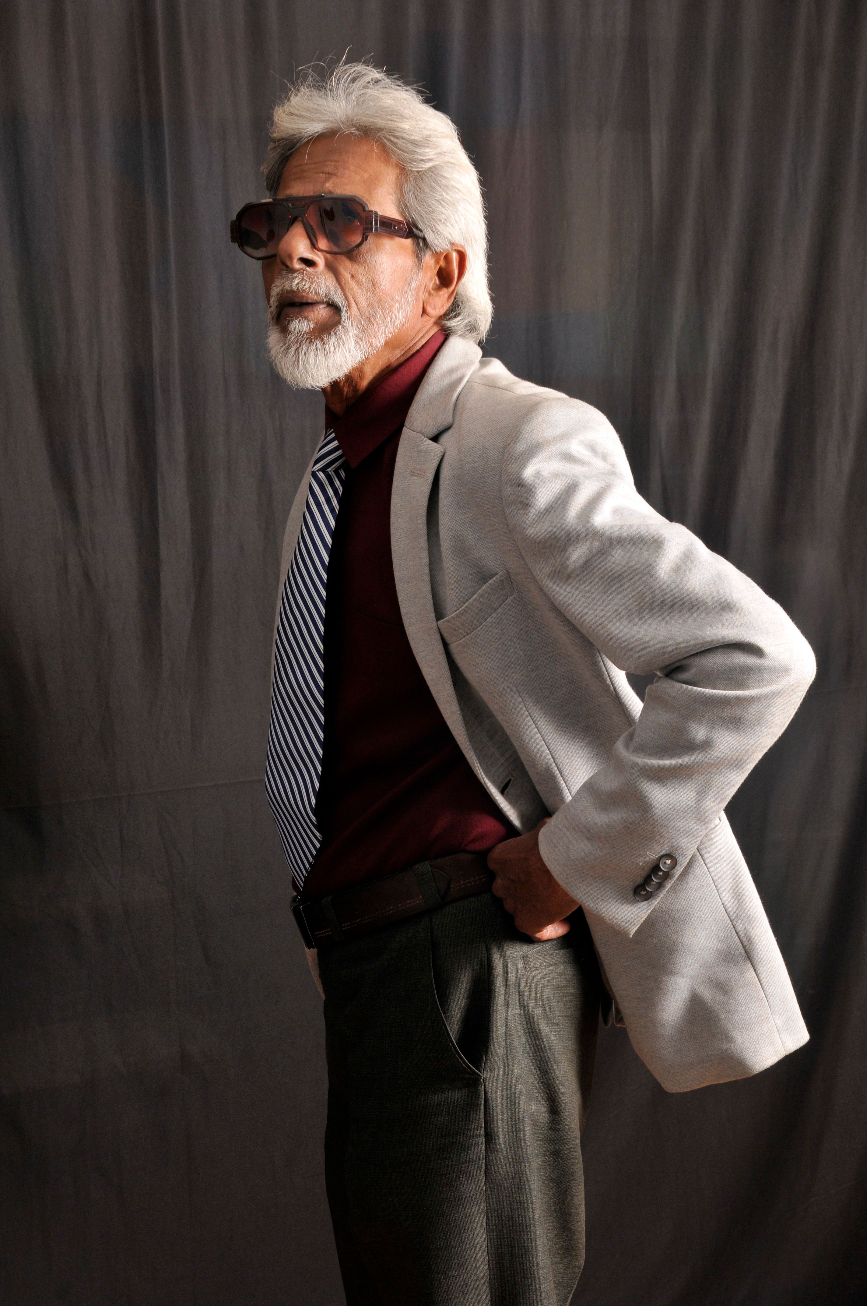 Confident senior man in a grey suit and sunglasses posing against a dark backdrop.