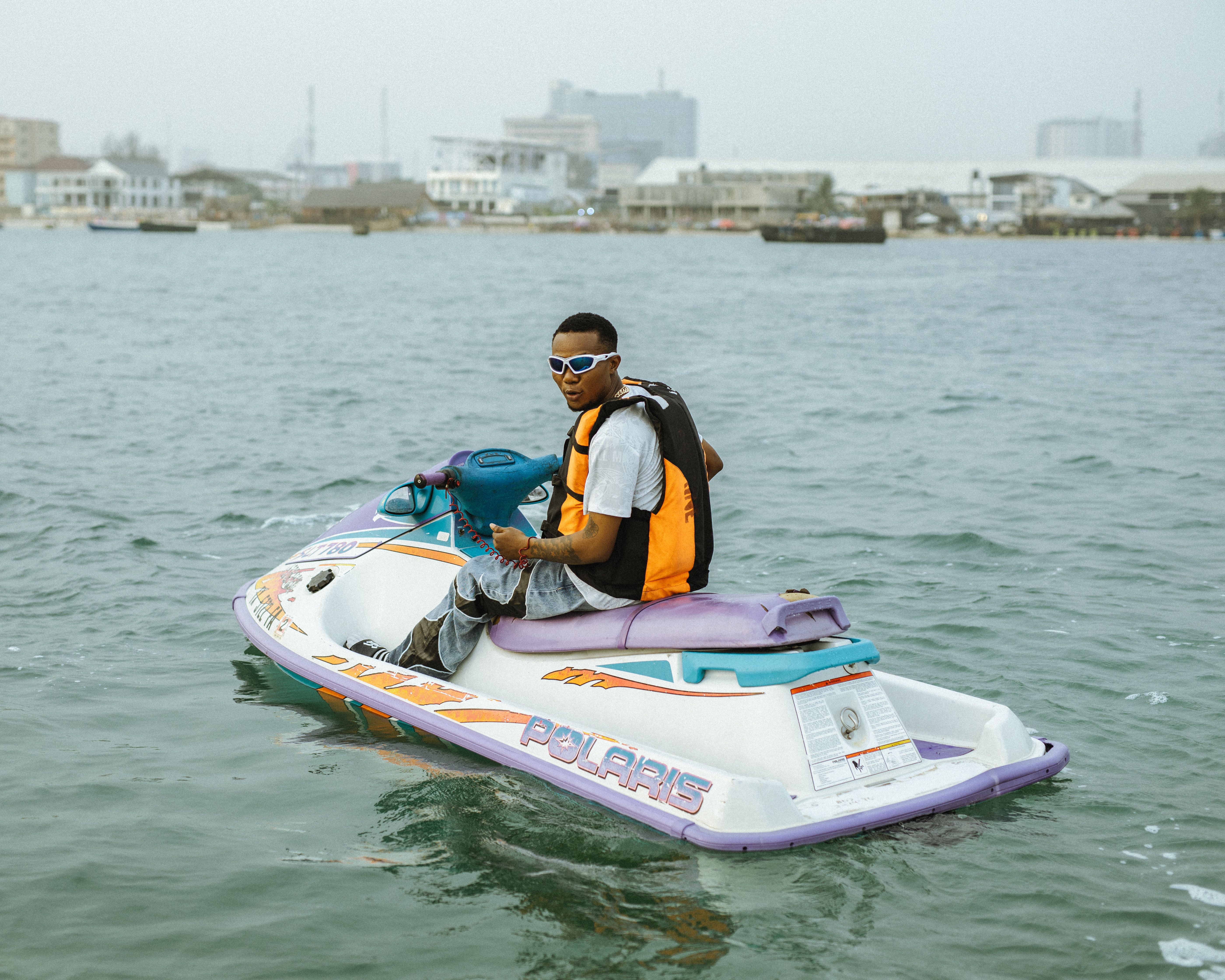 A man wearing a life jacket rides a jet ski on a river with an urban background.