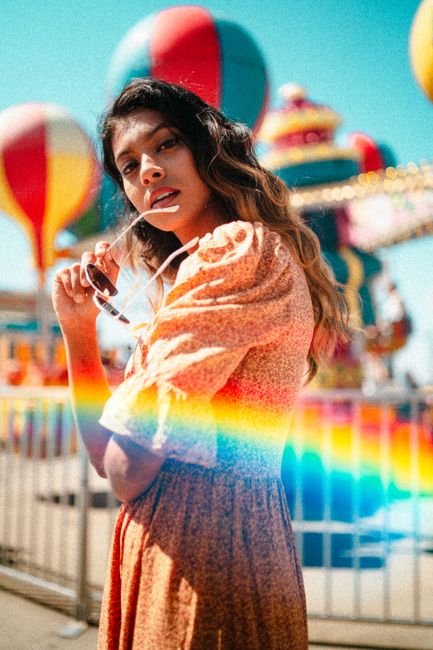 Stylish woman poses with sunglasses at colorful amusement park, capturing a summery fashion vibe.