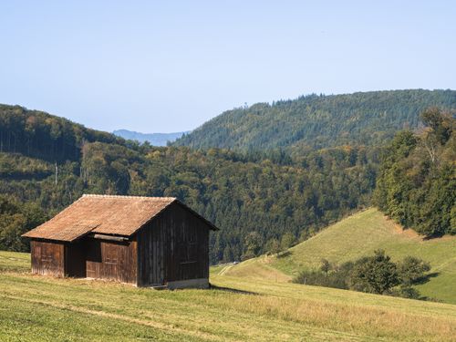 Free A solitary wooden barn in a serene, hilly landscape amidst lush greenery under a clear sky. Stock Photo